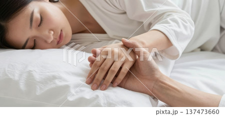 Close-up of a young Asian couple affectionately holding hands while the woman sleeps peacefully in a comfortable bed with white sheets. 137473660