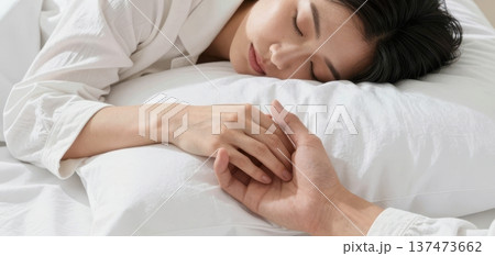 Close-up of a young Asian couple affectionately holding hands while the woman sleeps peacefully in a comfortable bed with white sheets. 137473662