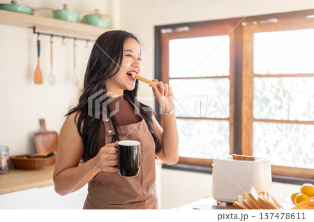 Pretty asian woman holding coffee mug enjoy eating a bread aside toaster on kitchen cooking counter. Pretty asian woman holding coffee mug enjoy eating a bread aside toaster on kitchen cooking counter. 137478611