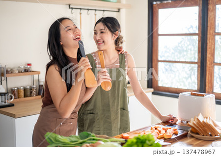 Asian woman couple girlfriends holding spatula singing at kitchen cook counter with food ingredients Asian woman couple girlfriends holding spatula singing at kitchen cook counter with food ingredients 137478967