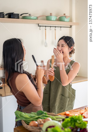Asian woman laughing at girlfriends holding spatula singing at kitchen counter while cooking a food. Asian woman laughing at girlfriends holding spatula singing at kitchen counter while cooking a food. 137478973