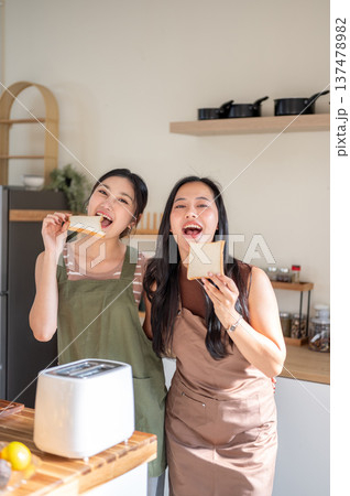 Pretty asian woman girlfriends holding a bread enjoy eating aside toaster on kitchen cooking counter Pretty asian woman girlfriends holding a bread enjoy eating aside toaster on kitchen cooking counter 137478982