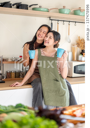 Smiling asian woman girlfriends holding couple coffee mugs while touching heads at kitchen counter. Smiling asian woman girlfriends holding couple coffee mugs while touching heads at kitchen counter. 137478996