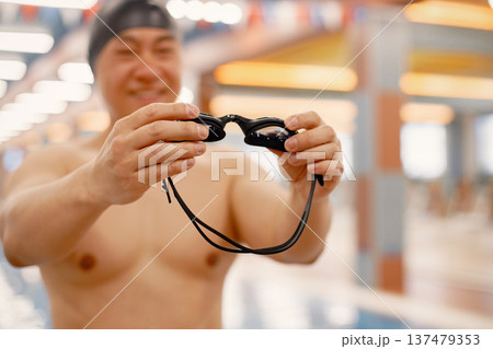 Focus on a goggles in man's hands. Man standing in in swimming pool. Professionale male swimmer wearing a hat. Focus on a goggles in man's hands. Man standing in in swimming pool. Professionale male swimmer wearing a hat. 137479353