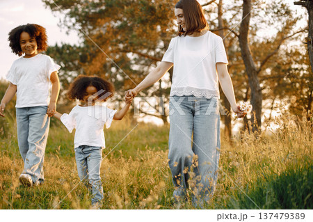 Photo of caucasian mother and two her african american daughters walking together outdoors. Girls and mother holding each others hands. Mother and daughers wearing white t-shirts. Photo of caucasian mother and two her african american daughters walking together outdoors. Girls and mother holding each others hands. Mother and daughers wearing white t-shirts. 137479389