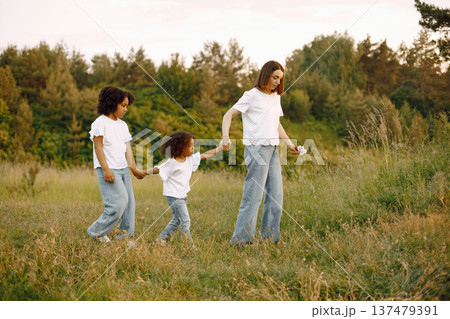 Photo of caucasian mother and two her african american daughters walking together outdoors. Girls and mother holding each others hands. Mother and daughers wearing white t-shirts. Photo of caucasian mother and two her african american daughters walking together outdoors. Girls and mother holding each others hands. Mother and daughers wearing white t-shirts. 137479391