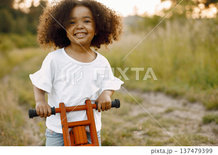 Happy toddler girl riding red bike. Little kid learns to ride a bicycle. African girl has black curly hair. Happy toddler girl riding red bike. Little kid learns to ride a bicycle. African girl has black curly hair. 137479399