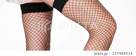 Close-up of female legs in black fishnet stockings against clean white background. Elegant fashion apparel and soft lighting emphasize fashion, sensuality Close-up of female legs in black fishnet stockings against clean white background. Elegant fashion apparel and soft lighting emphasize fashion, sensuality 137484514