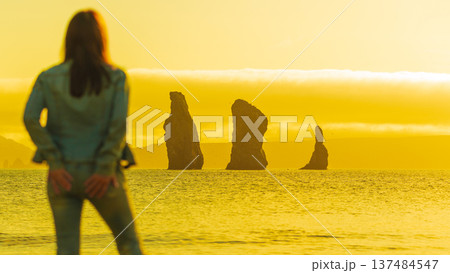A blurred woman from behind admires a serene ocean view with silhouetted sea stacks. The entire landscape is drenched in a golden yellow light, evoking a peaceful, travel mood. Focus on background A blurred woman from behind admires a serene ocean view with silhouetted sea stacks. The entire landscape is drenched in a golden yellow light, evoking a peaceful, travel mood. Focus on background 137484547