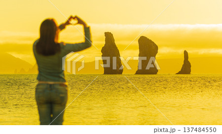 A woman from behind makes a heart shape with her hands, framing sea stacks in the ocean. The scene is bathed in a warm, golden sunset light, creating a romantic mood. Selective focus on background 137484550