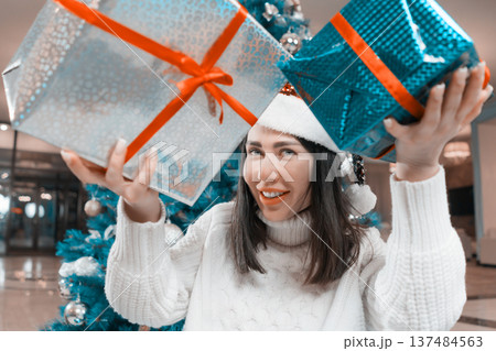 A joyful woman in a white sweater and Santa hat smiles while holding two large Xmas presents. Festive holiday scene with a decorated Christmas tree. Modern teal, red and silver color palette A joyful woman in a white sweater and Santa hat smiles while holding two large Xmas presents. Festive holiday scene with a decorated Christmas tree. Modern teal, red and silver color palette 137484563