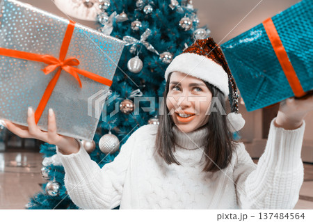 A happy young woman in a white sweater and sparkly red Santa hat smiles holding two Xmas gift boxes. A decorated blue Christmas tree with silver ornaments is in the background. Festive holiday mood 137484564