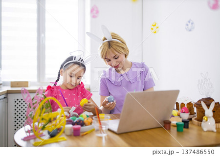 A mother and daughter wearing bunny ears paint Easter eggs together at a table while looking at a laptop screen, celebrating the holiday 137486550