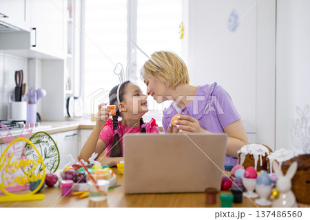 A mother and daughter wearing bunny ears share a tender moment while decorating Easter eggs together in a bright kitchen 137486560