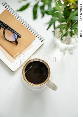 A cup of coffee, notepads, glasses, and a vase of flowers on a white table. A cup of coffee, notepads, glasses, and a vase of flowers on a white table. 137486634