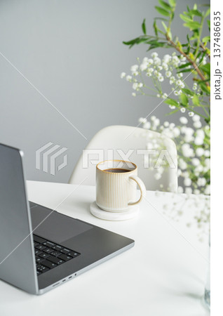 A black cup of coffee, laptop on a white table with gypsophila flowers in a glass vase on a gray background. A black cup of coffee, laptop on a white table with gypsophila flowers in a glass vase on a gray background. 137486635