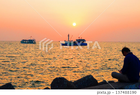 Sunset at shore with fishing boats. Person sitting on pier by sunset sea. Sunset over ocean 137488190