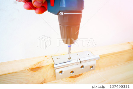 Screwdriver is used to attach hinge to wooden door frame. Cordless drill to secure silver hinge Screwdriver is used to attach hinge to wooden door frame. Cordless drill to secure silver hinge 137490331