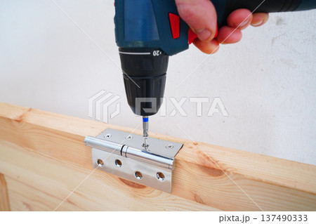 Screwdriver is used to attach hinge to wooden door frame. Cordless drill to secure silver hinge Screwdriver is used to attach hinge to wooden door frame. Cordless drill to secure silver hinge 137490333