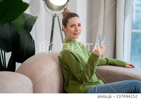 Pretty young woman in green sitting on the sofa with a glass of water Pretty young woman in green sitting on the sofa with a glass of water 137494252