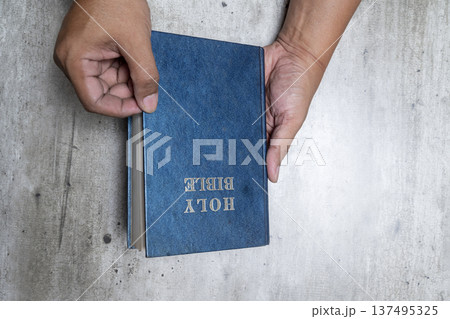 A top-down view of hands holding and starting to open a blue leather-bound Holy Bible with gold lettering on a rustic, textured gray surface. Captures themes of faith, study, and devotion A top-down view of hands holding and starting to open a blue leather-bound Holy Bible with gold lettering on a rustic, textured gray surface. Captures themes of faith, study, and devotion 137495325