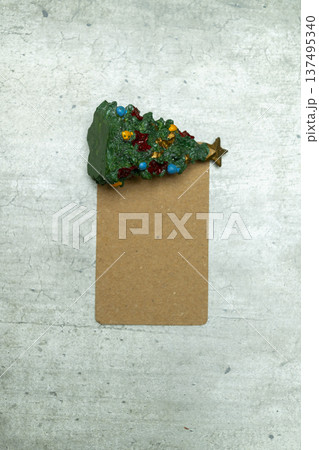 A festive green miniature Christmas tree decoration lying on a blank brown cardboard gift tag over a textured gray stone-like surface. Perfect for holiday greetings and labels 137495340