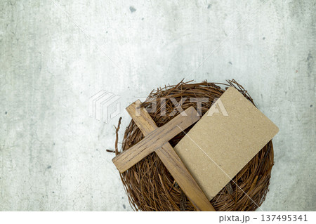 A wooden Christian cross and blank craft paper card in a twig nest on a grey textured surface, suitable for religious themes, Easter greetings, or sympathy cards 137495341
