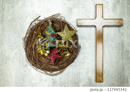 Top view of a wooden cross next to a bird's nest filled with Christmas tree ornaments, stars, and bells on a textured gray background 137495342