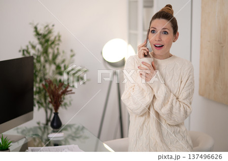 Woman in white sweater talking on the phone and looking surprised 137496626