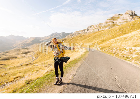 Hitchhiking woman traveler try to stop car on the mountain road. Travel and vacation theme 137496711