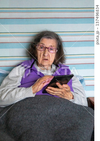 Elderly woman in bed using a tablet at home, wearing glasses and a purple vest 137496834