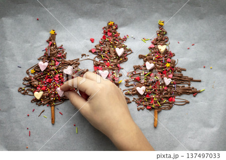 Hand decorating chocolate Christmas tree cookies with colorful sprinkles and heart decorations for festive dessert 137497033