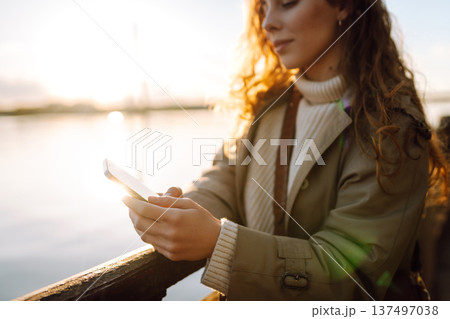Young female city dweller with mobile phone standing on embankment alone. Woman shopping online. Young female city dweller with mobile phone standing on embankment alone. Woman shopping online. 137497038
