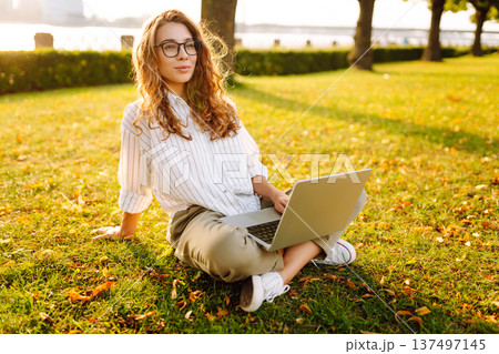 Young Woman sitting on grass ground, working on laptop. Freelance work, technology concept. Young Woman sitting on grass ground, working on laptop. Freelance work, technology concept. 137497145