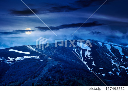 mysterious nature image of ridge with snow capped tops in spring landscape. mountain background wallpaper at night. full moon over greenery scenery with forested slopes under sky with clouds 137498282