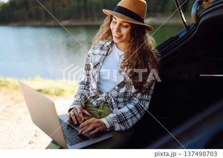 Woman sits on trunk of car, using her laptop while enjoying tranquility of a lake. Remote work. 137498703