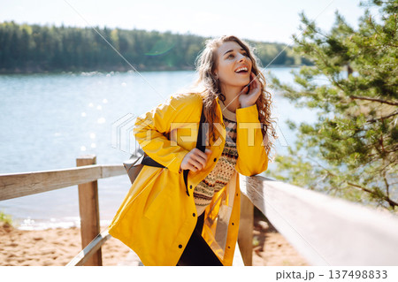 A cheerful young woman in a yellow jacket enjoying a sunny day by the lake. Trips. Active lifestyle. 137498833