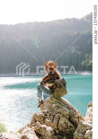 Back view young woman with backpack looking the view of the mountain lake in sunny weather. Back view young woman with backpack looking the view of the mountain lake in sunny weather. 137499205