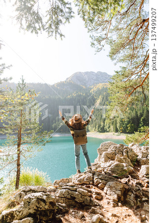 Back view young woman with backpack looking the view of the mountain lake in sunny weather. Back view young woman with backpack looking the view of the mountain lake in sunny weather. 137499207