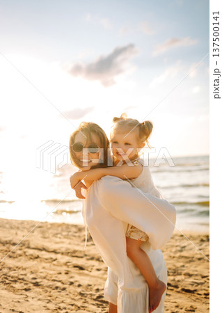 A mother and daughter enjoy a sunny beach stroll, as waves gently lap at their feet during sunset 137500141