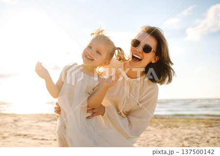 A mother and daughter enjoy a sunny beach stroll, as waves gently lap at their feet during sunset A mother and daughter enjoy a sunny beach stroll, as waves gently lap at their feet during sunset 137500142