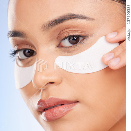 Beauty, dermatology and portrait of woman with collagen patch for anti aging or skin glow on blue background. Cosmetics, facial mask and face of model with skincare product for dermatology in studio Beauty, dermatology and portrait of woman with collagen patch for anti aging or skin glow on blue background. Cosmetics, facial mask and face of model with skincare product for dermatology in studio 137500588