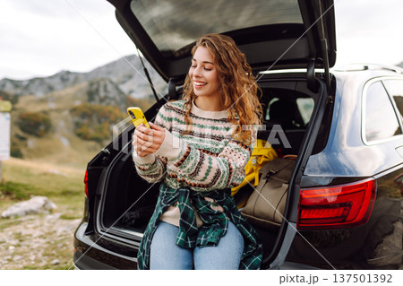Young traveler woman sits on the rear of her car, smiling and using phone in scenic mountainous area 137501392