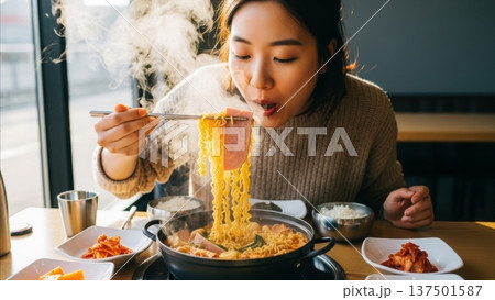 Young Asian woman eating steaming hot ramen noodles with chopsticks in cozy restaurant setting with kimchi side dishes 137501587