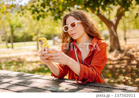 Young woman sits on bench in park, using smartphone while dressed casually. Travel, blogging concept Young woman sits on bench in park, using smartphone while dressed casually. Travel, blogging concept 137502011