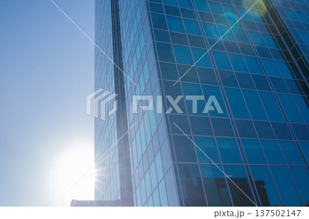 City skyscraper building exterior facade with blue sky background and bright sun shining behind the building. Repetitive windows made of reflective glass. Big city life, contemporary architecture City skyscraper building exterior facade with blue sky background and bright sun shining behind the building. Repetitive windows made of reflective glass. Big city life, contemporary architecture 137502147
