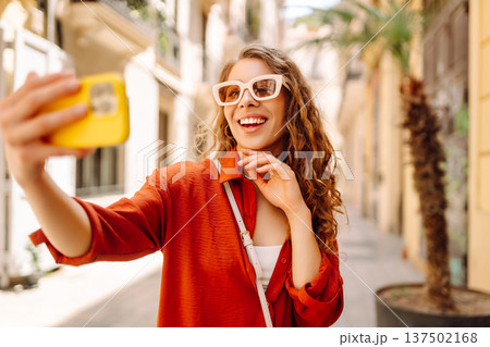 Cheerful woman taking a selfie in stylish sunglasses while strolling down a sunlit city street Cheerful woman taking a selfie in stylish sunglasses while strolling down a sunlit city street 137502168