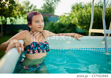 Teen girl enjoying a summer afternoon in a backyard pool surrounded by lush greenery and sunshine 137502668