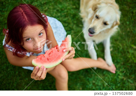 Teen girl enjoying watermelon while sitting on grass with a dog nearby during a sunny day in the backyard 137502680