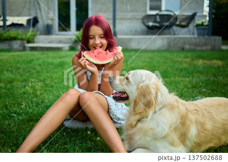 Teen girl enjoying watermelon while sitting on grass with a dog nearby during a sunny day in the backyard Teen girl enjoying watermelon while sitting on grass with a dog nearby during a sunny day in the backyard 137502688